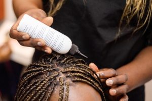 A hair stylist applies hair product from a bottle to the scalp of a woman with braids.