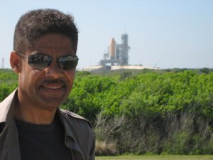 Ken Phillips, wearing sunglasses, with a space shuttle on the launching pad behind him in the distance.