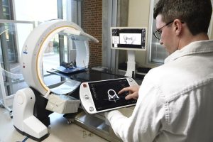 Benjamin Killeen, a PhD student at Johns Hopkins, looks at a 3D image on his tablet. He is standing next to a Loop-X medical imagine robot, a ring-shaped device that has an operating table under it.