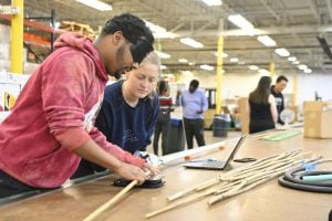 A student and an associate at Blind Industries and Services stand at a table working on assembling a cane.