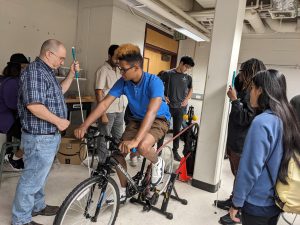Stephen Belkoff, associate research professor, and students from the BISM gather around a bike hooked up to a governor. There is a student pedaling the bike.