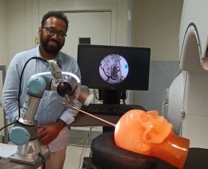 Prasad Vagdargi, smiling, next to a robotic surgical device demonstrating a brain operation technique on a clear mannikin head.