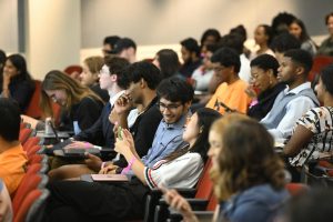 A student shares their smartphone screen with a student sitting besides them among a group of students seated in auditorium seating.