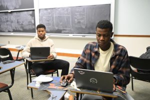Two students work on laptops at desks in front of a blackboard.
