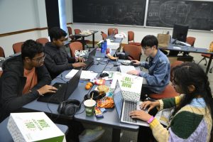 4 students sit around a table working on laptops. The middle of the table has various junk food on it.