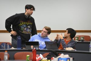 A student stands, talking with two seated students working at a desk.