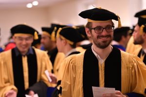 Man with glasses and beard is smiling; he is wearing a doctoral graduation cap and gown and is surrounded by other doctoral graduates. 
