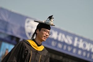 Man with graduation cap and gown stands in front of stage. He is smiling and has a small stuffed animal bluejay on his cap; the bluejay has it's own small graduation cap.
