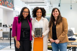 Alexis Pena poses for a photo, holding a plaque. On her right is Dr. Darlene Saporu and on her left, Professor Rebecca Shulman. 