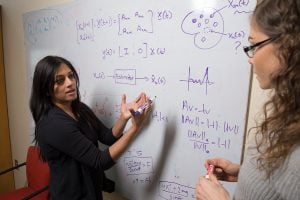 Professor Sri Sarma writes an equation on a dry erase board while talking to a student.