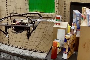 A quadcopter uses an outstretched arm to grasp a can of tomato soup from a pile of groceries.