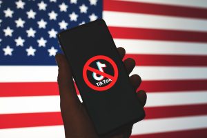 A hand holds a phone with the red X through the TikTok logo. An American flag is in the background.