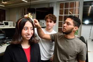 In an electronics lab, Christian Kanzki, and Kyle Cooper, adjust a device mounted to a headband on Rita Rozental's head.