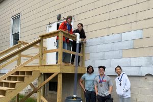 A team of students stands alongside a building. Two are on an eleveated porch. A tube runs from a trash can on porch to one on the ground.