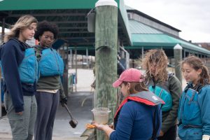 Four students on a dock and boat gather around an instructor holding a bucket of water with oysters. 