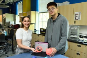 Allison Shannon and Zhehao Zhang stand at a table in a lab holding their prototype device, with a laptop on the table. 