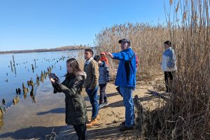Students and a faculty member observe the bay from a marsh area on the shore. One student is taking a photo of something out-of-frame.