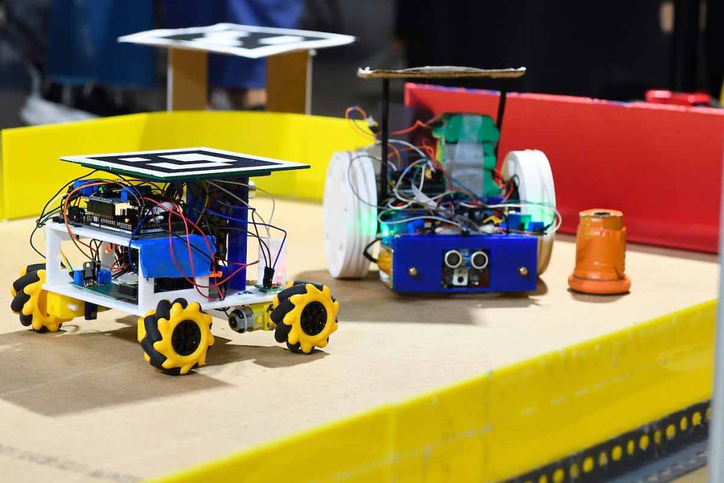 Two small robots face on a mini hockey rink, trying to push a "puck" that resembles a small traffic cone. 