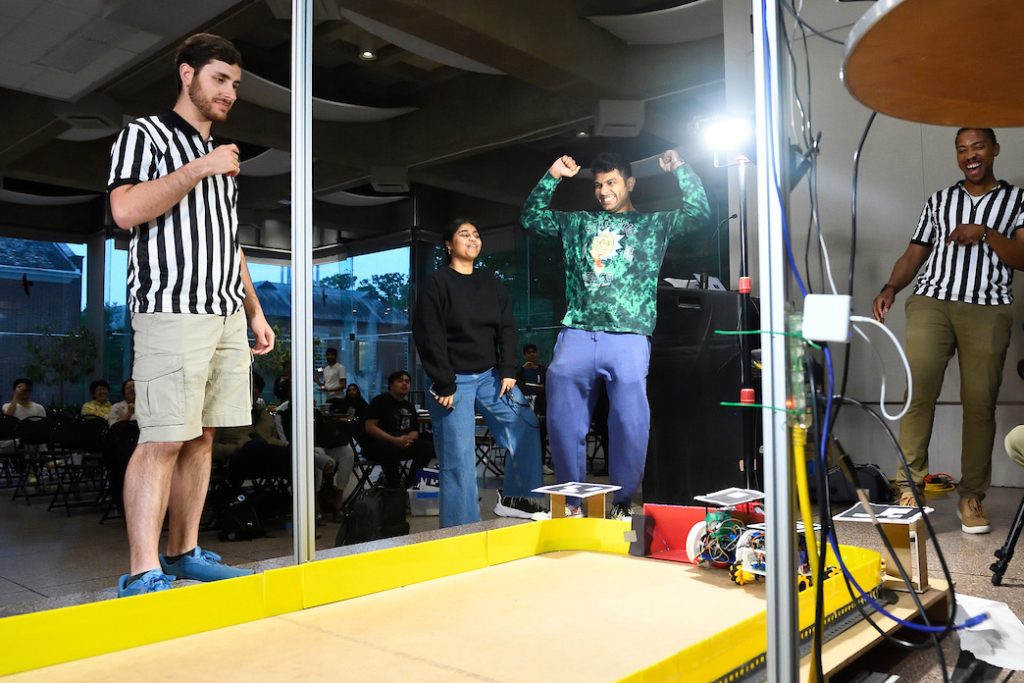 A student raises his arms as a small robot puts a "puck" in a goal on a simulated hockey rink. A ref, teammate, and Professor Jeremy Brown (laughing) look on. 