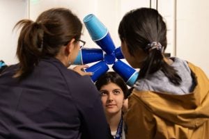 Two students assemble a crown of blue plastic cups on a fellow student. 