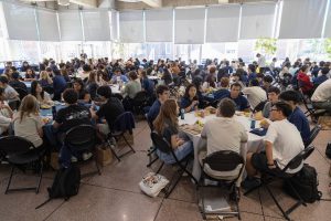 The Glass Pavilion, filled with tables, surrounded by students and faculty members having lunch and chatting. 