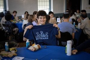 A student sits at a round table holding up a "Meet the Flock" blue shirt. 