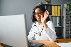Photo of smiling female doctor waving toward computer screen.