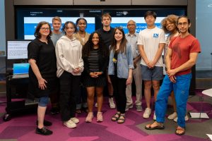 11 students and faculty members pose for a group photo in front of two large screens.