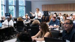 This image depicts a Faculty Forward collaborative workshop in a modern conference room. The room is filled with attendees seated at round tables, engaging in discussions and taking notes. A facilitator, standing in the center of the room, gestures as she addresses the group, holding a tablet or phone. The room has large windows allowing natural light to enter, and a refreshment station with coffee and snacks is visible in the background. Laptops and notebooks are scattered across the tables, indicating a working session. 