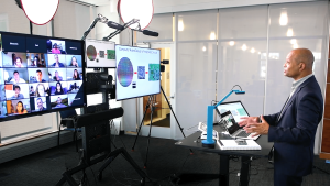 This image shows a person standing and giving a lecture in a professional video production setup. Multiple large monitors display a Zoom call with participants in a grid layout and a slide presentation on integrated circuits. The speaker, wearing a suit, is positioned at a podium with a laptop, a tablet, and a small document camera. Professional studio lighting and audio equipment, including overhead lights and a mounted microphone, are set up to ensure clear visibility and sound. The background consists of glass panels and a modern office-like environment.