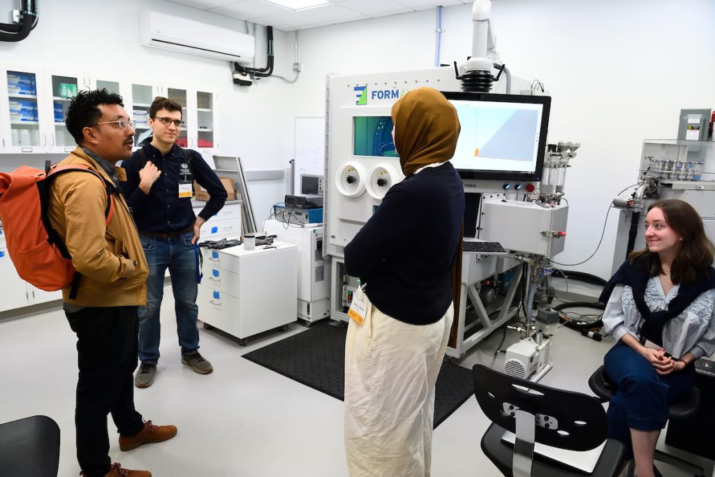 A tour guide chats with attendees in a lab filled with materials characterization equipment.