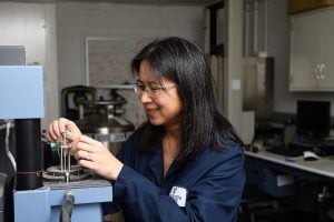 Vicky Nguyen adjusts a precision instrument in a laboratory, focusing intently on the setup. Scientific equipment and a whiteboard with equations are visible in the background.