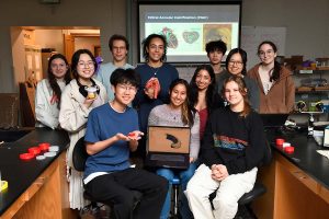 Group photo of team around a laptop. One student holds the value prototype. 