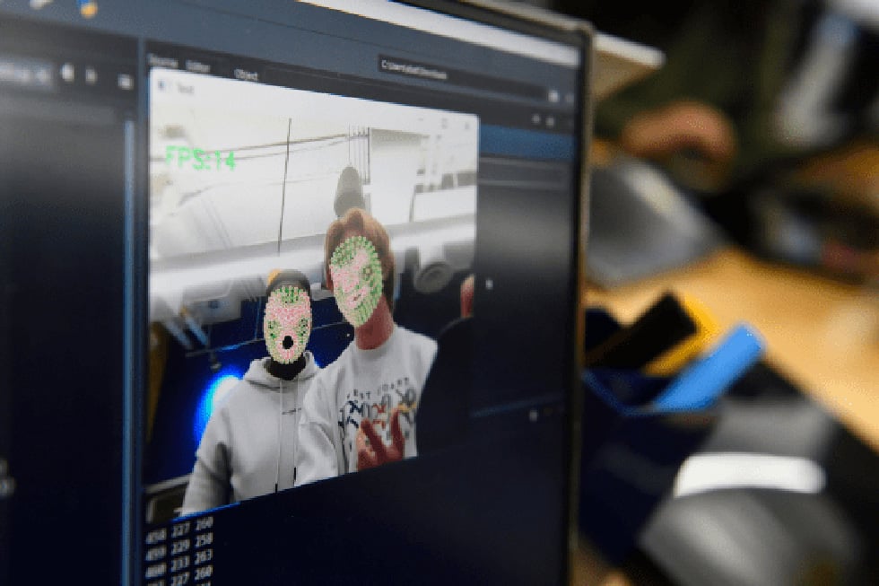 Students look at themselves on a computer screen with their faces overlayed by face-mapping software. 