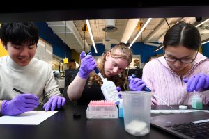 Three students using pipettes and lab equipment at a black table.