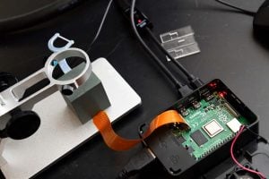 Close-up of a Raspberry Pi connected to a camera module and microscope stand, with slides visible in the background