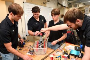 The four members of the team work on their prototype on a table filled with spools of wire, tools and a very full breadboard connected to the machine.