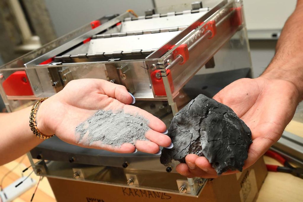 Two hands show an asteroid-like rock compared with fine powder. A grinding device is in the background. 