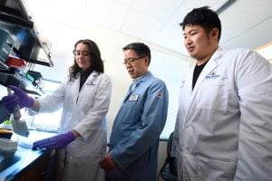 Professor Hai-Quan Mao observes a lab member using a pipette while another team member looks on, all wearing lab coats in a research lab.