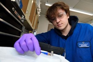 Liam McMullin conducts a lab experiment wearing safety glasses and gloves, part of his work optimizing cathode materials.