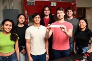 Seven students pose in a lab, one holding a prototype of plant-based drywall.