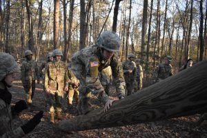 Jenna Halpin, in full Army fatigues and helmet, climbs over a large log obstacle as fellow ROTC cadets look on in a wooded training area.