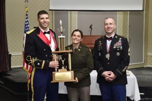 Jenna Halpin stands between two uniformed officers, smiling and holding a large trophy at an ROTC awards ceremony.