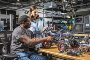A younger woman is standing and a man is seated beside her; both are looking at and tinkering with a robot. The robot is a small, 15 inch object that looks like a simple vehicle with wheels. 