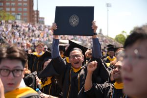 Joyful graduate holding up diploma cover, surrounded by fellow graduates and crowd