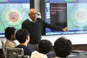 Professor K.T. Ramesh leads a class seated around a conference table. Behind him, screens show concentric circles of a theoretical asteroid impact zone in Africa. 