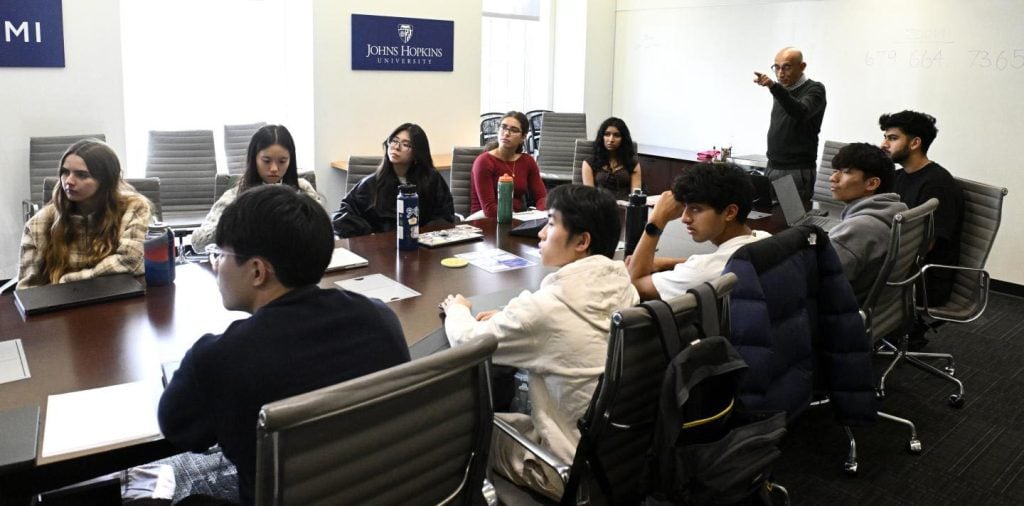 K.T. Ramesh points towards a screen (off-camera) in front of a class of students at a conference table.