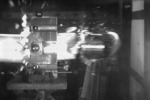 Black-and-white high-speed image of a laboratory impact experiment, showing a metal plate colliding with a target apparatus and producing a bright flash at the point of contact.