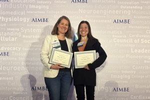 Hicks and Spangler pose for a photo together, holding their recognition certificates against an AIMBE backdrop. 
