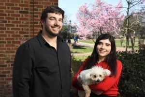 Eric Nalisnick and Alesso Carbo pose for a photo outdoors on campus with cherry blossoms in the background. Carbo is holding her dog. 